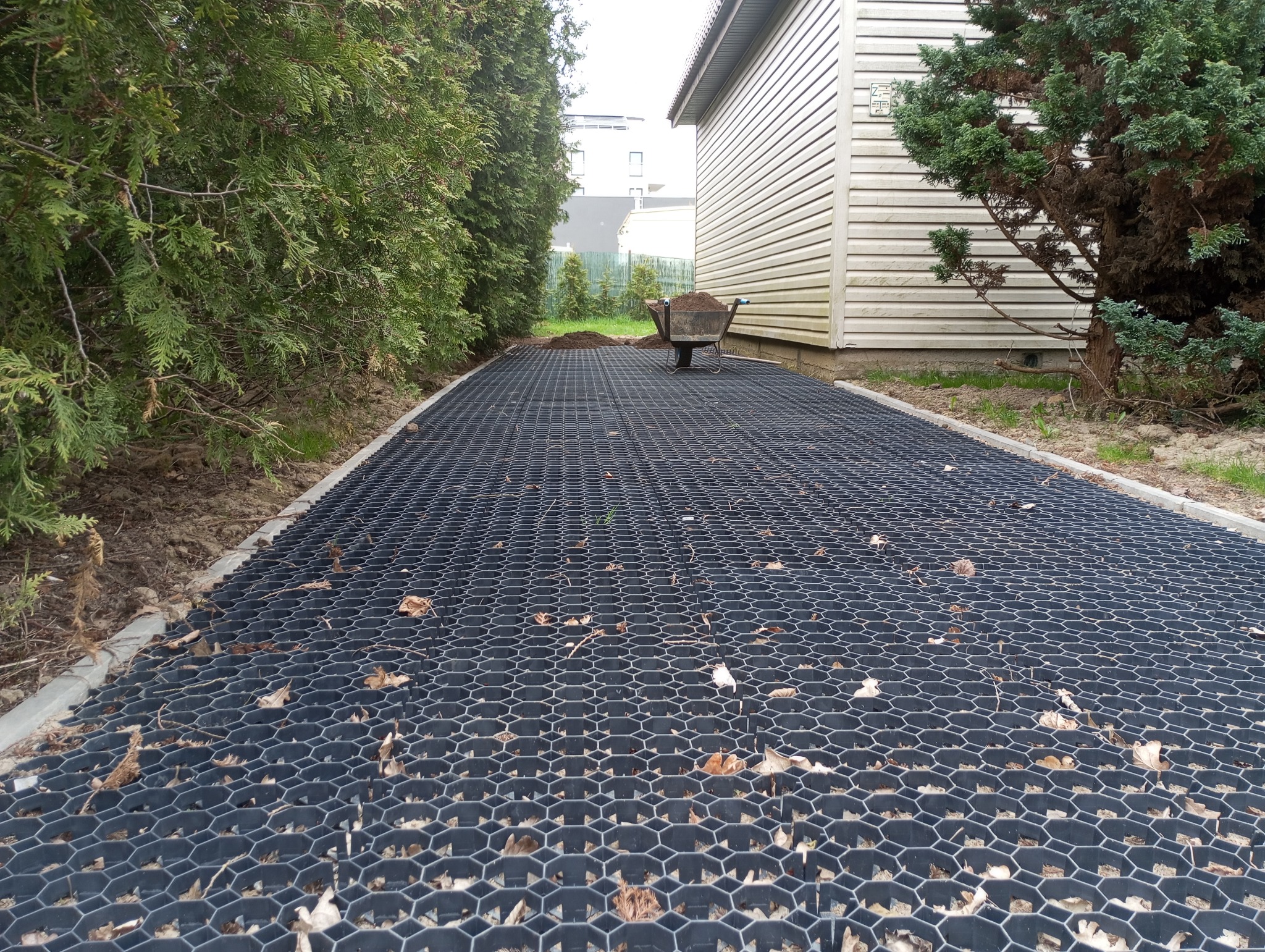 Czarna, ażurowa nawierzchnia z obrzeżami, prawdopodobnie przygotowana pod żwir lub kamień, z taczką ziemi w tle, otoczona zielenią.