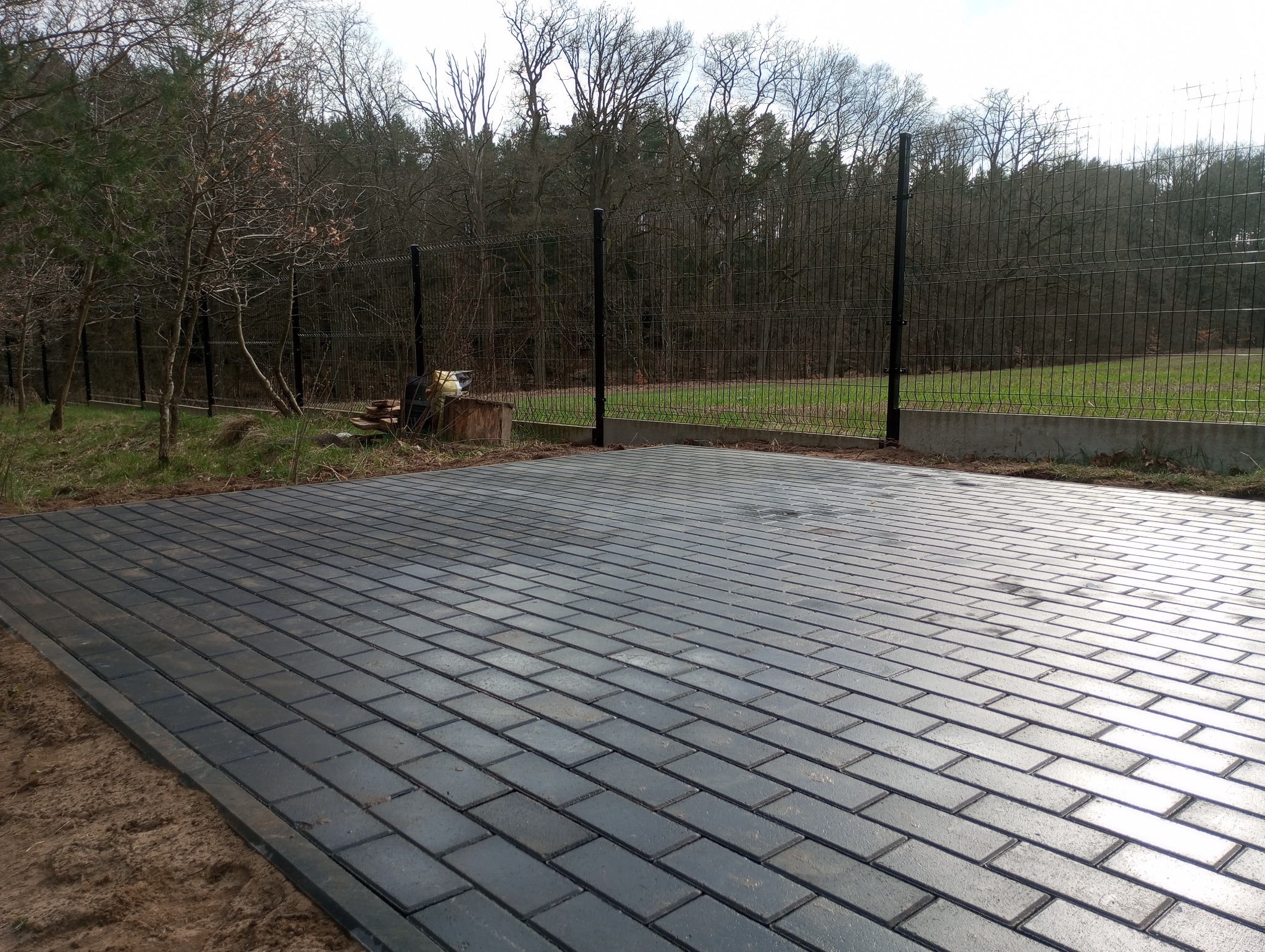 Ułożona, ciemnoszara kostka brukowa w kształcie prostokątów, obrzeża z piasku, w tle siatka ogrodzeniowa i drzewa bez liści. Widok z perspektywy.