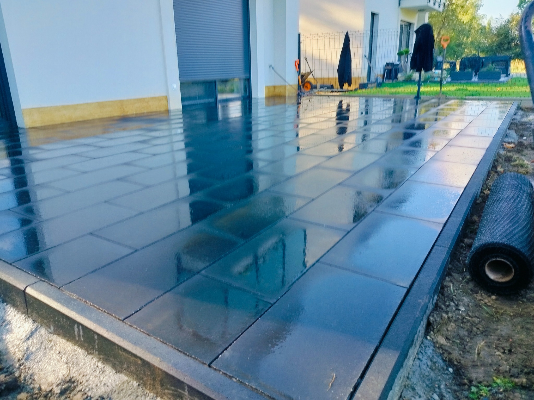 Świeżo ułożona, mokra kostka brukowa w odcieniach szarości, odbijająca otoczenie. Widoczny fragment budynku z bramą garażową i rolka materiału geotekstylnego.