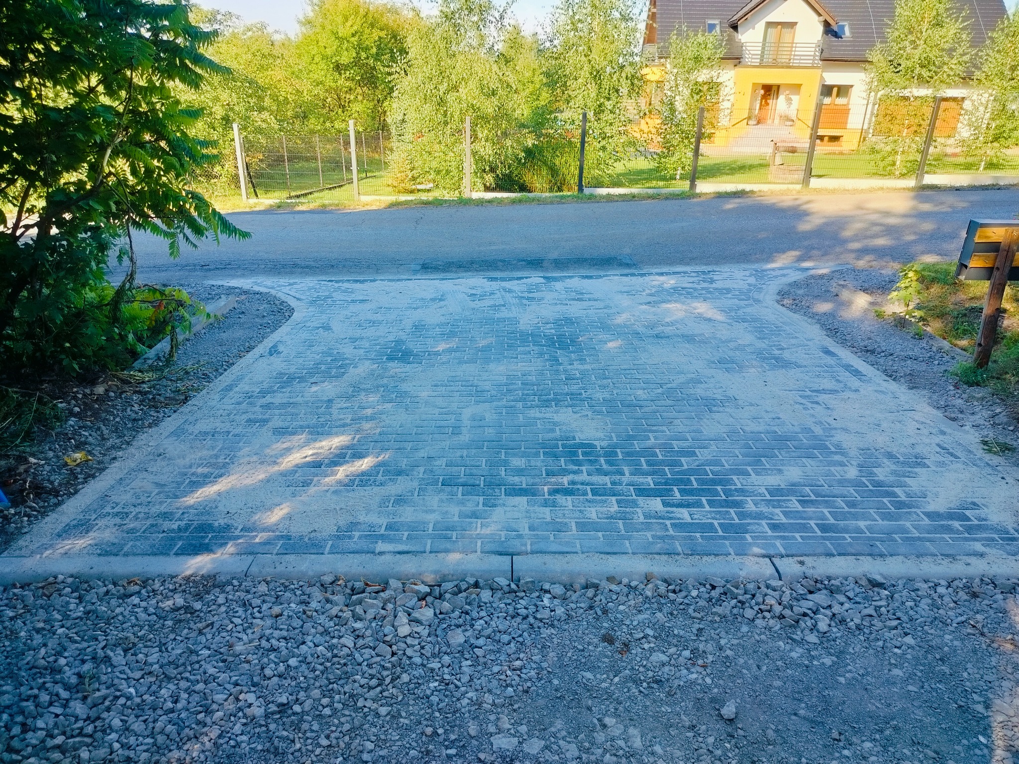 Ułożona kostka brukowa w odcieniach szarości, obrzeża z kamieni. Widok na dom z ogrodem w tle. Świeżo wykonana nawierzchnia.