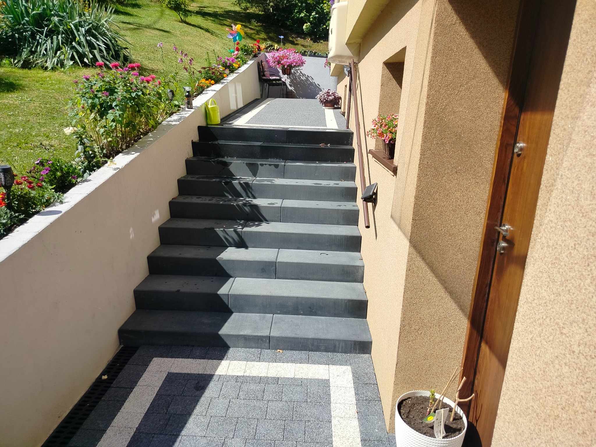 Schody z kostki brukowej prowadzące do domu, z widocznym ogrodem i kwiatami. Detale wykończenia, takie jak donice i kolorystyka kostki, nadają unikalny charakter.