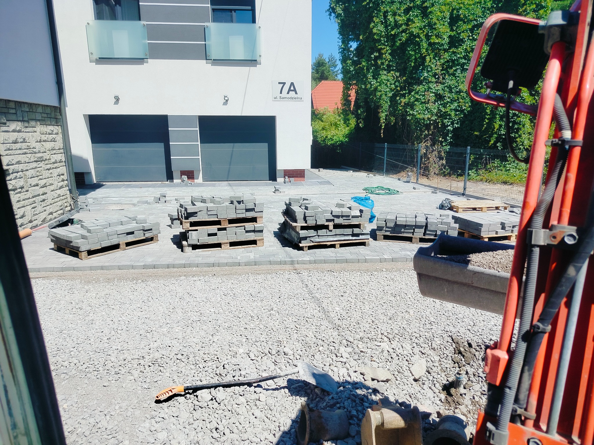 Widok z kabiny na plac budowy: palety z kostką brukową przed nowoczesnym domem z dwoma garażami, w tle fragment koparki i przygotowane podłoże.