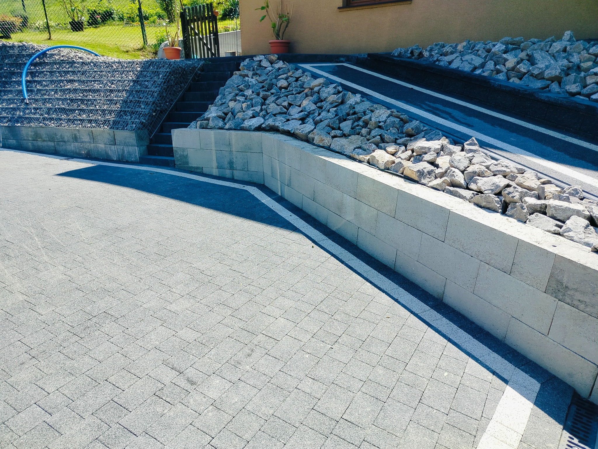 Ułożona kostka brukowa w szarym odcieniu. Widoczna ściana oporowa z kamieniami i schody. Elementy drenażu liniowego. Całość w jasnym, słonecznym oświetleniu.