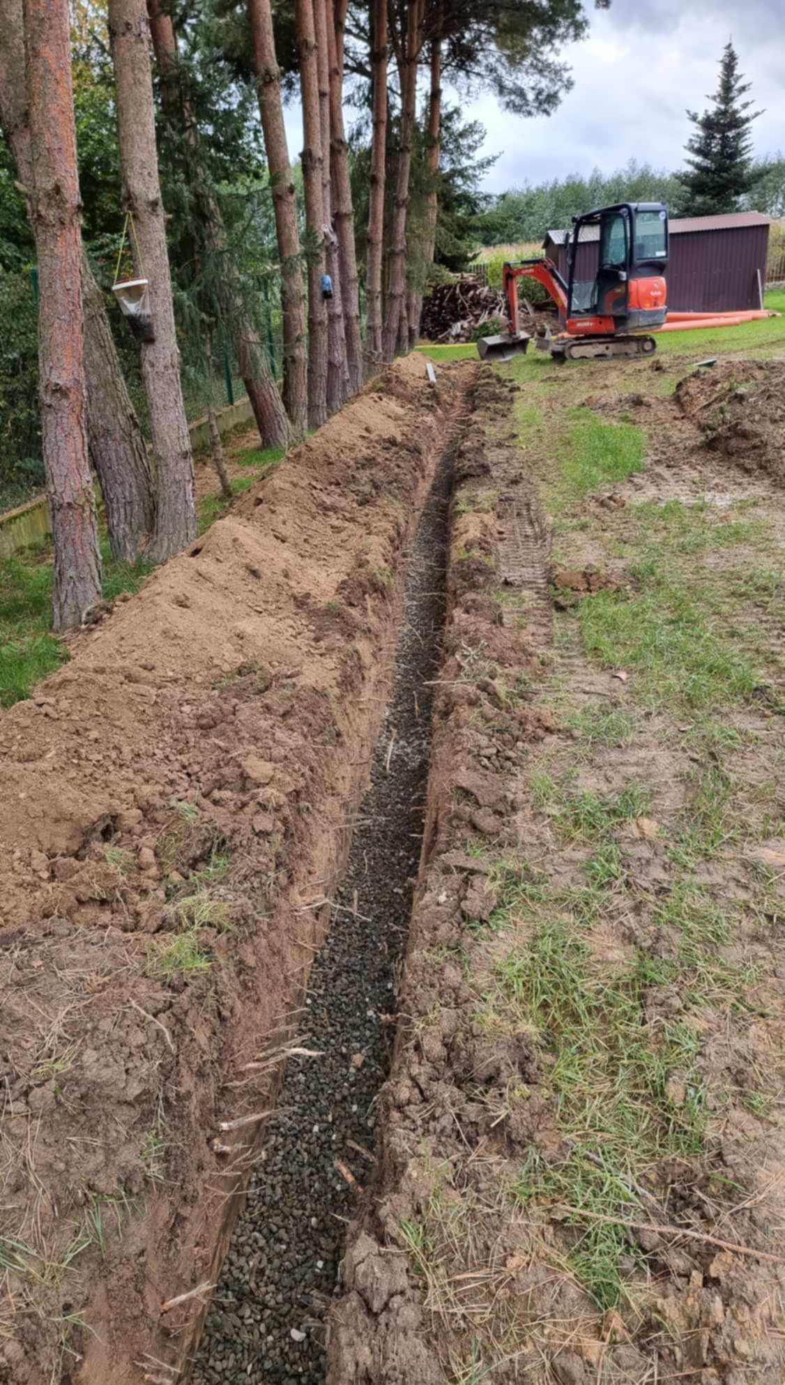 Wąski wykop wypełniony żwirem, ciągnący się wzdłuż rzędu drzew. W tle widoczna minikoparka i sterta drewna. Prace ziemne w ogrodzie.