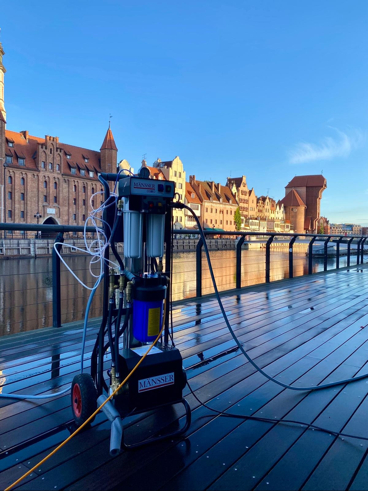 Profesjonalna maszyna do czyszczenia elewacji MÄNSSER na tle gdańskiej starówki, po umyciu mokrej drewnianej podłogi. Błękitne niebo.