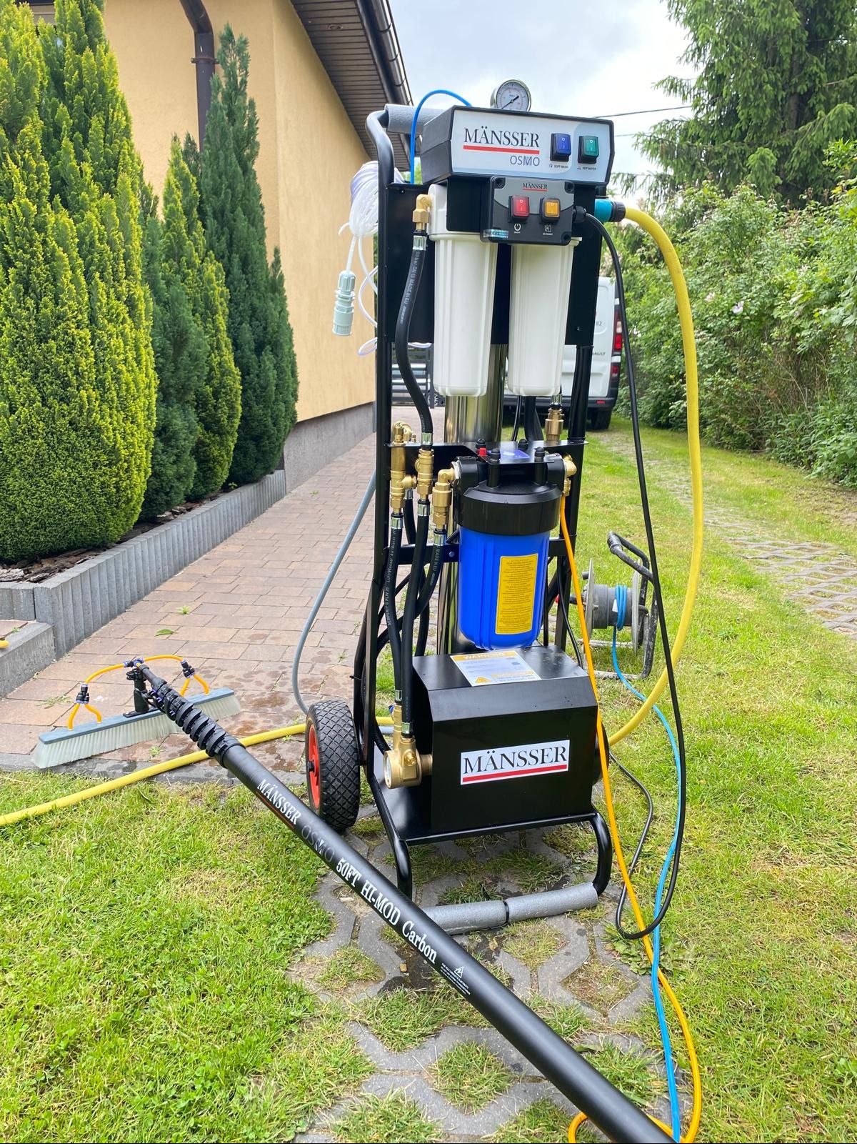 Mobilny system filtracji wody MÄNSSER OSMO z teleskopową lancą i szczotką na trawie, gotowy do mycia elewacji. Widoczny budynek w tle.
