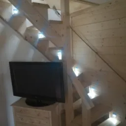 Drewniane schody z podświetleniem LED wkomponowane w drewnianą zabudowę, obok komoda z telewizorem, widoczna lina mocująca na ścianie.