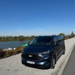 Niebieski Ford Transit Custom w Krakowie, zaparkowany na żwirowym parkingu z widokiem na jezioro w słoneczny dzień. Auto z przodu, tablica rejestracyjna widoczna.