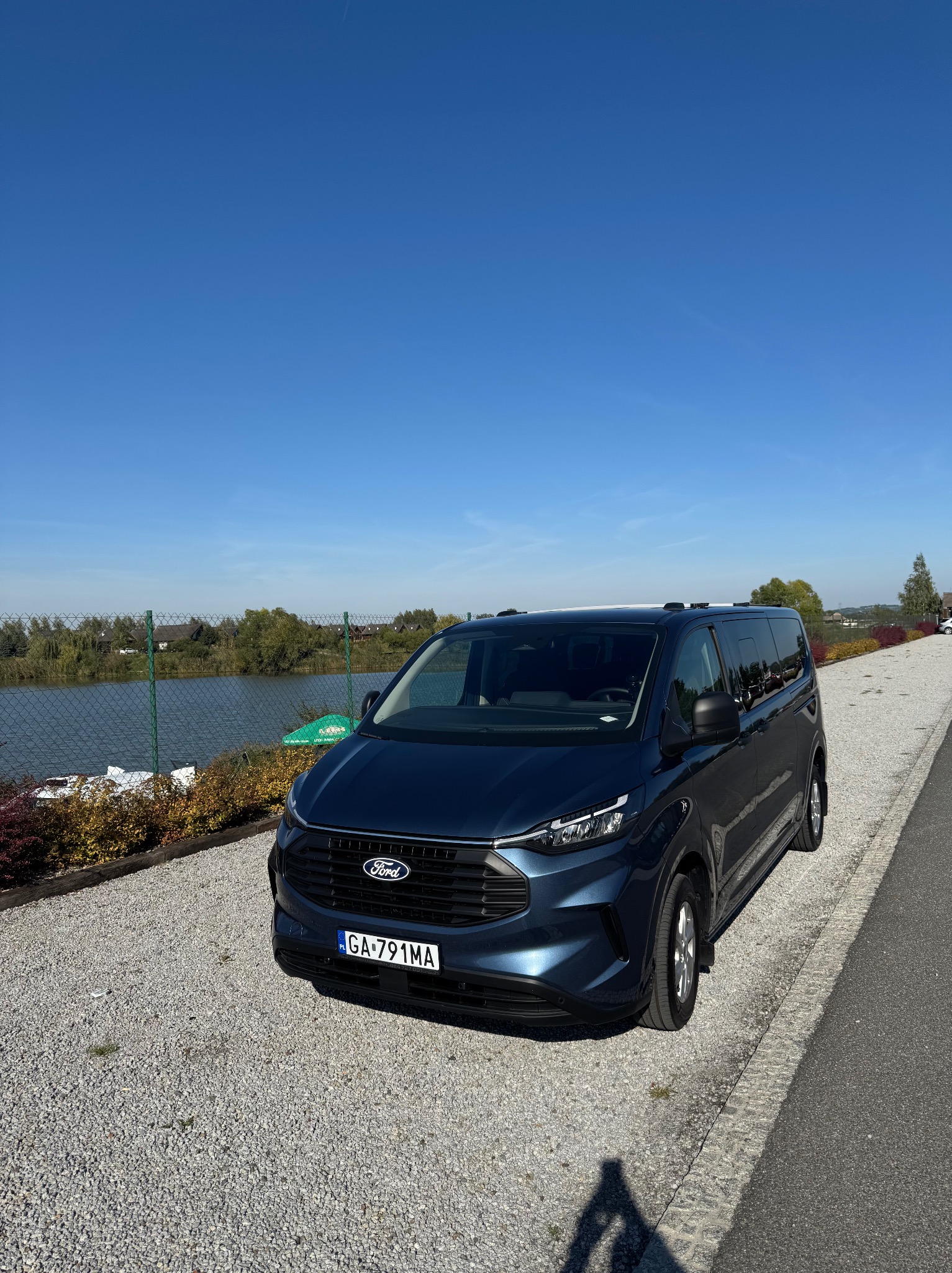 Niebieski Ford Transit Custom w Krakowie, zaparkowany na żwirowym parkingu z widokiem na jezioro w słoneczny dzień. Auto z przodu, tablica rejestracyjna widoczna.