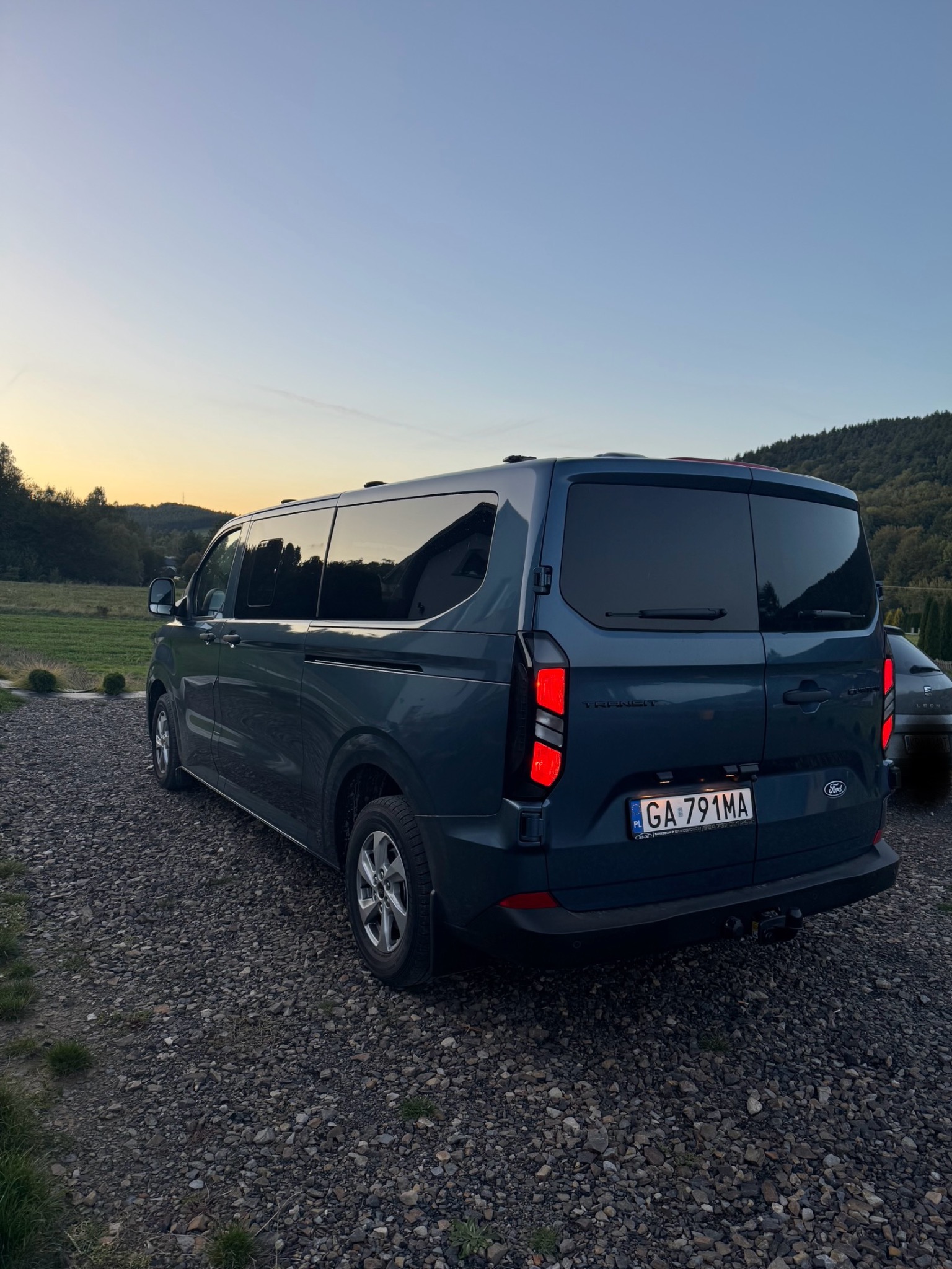 Niebieski Ford Transit z przyciemnianymi szybami, zaparkowany na żwirowym podłożu, z widocznymi tylnymi światłami i tablicą rejestracyjną, w tle zielone wzgórza i zachodzące słońce.