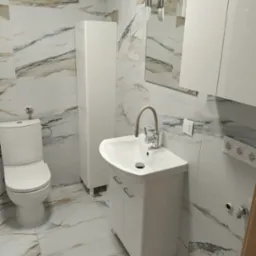 Wykończona łazienka w jasnych marmurkowych płytkach. Biała toaleta, szafka pod umywalką i słupek. Bateria z beżowym wężykiem. Lustro z oświetleniem.