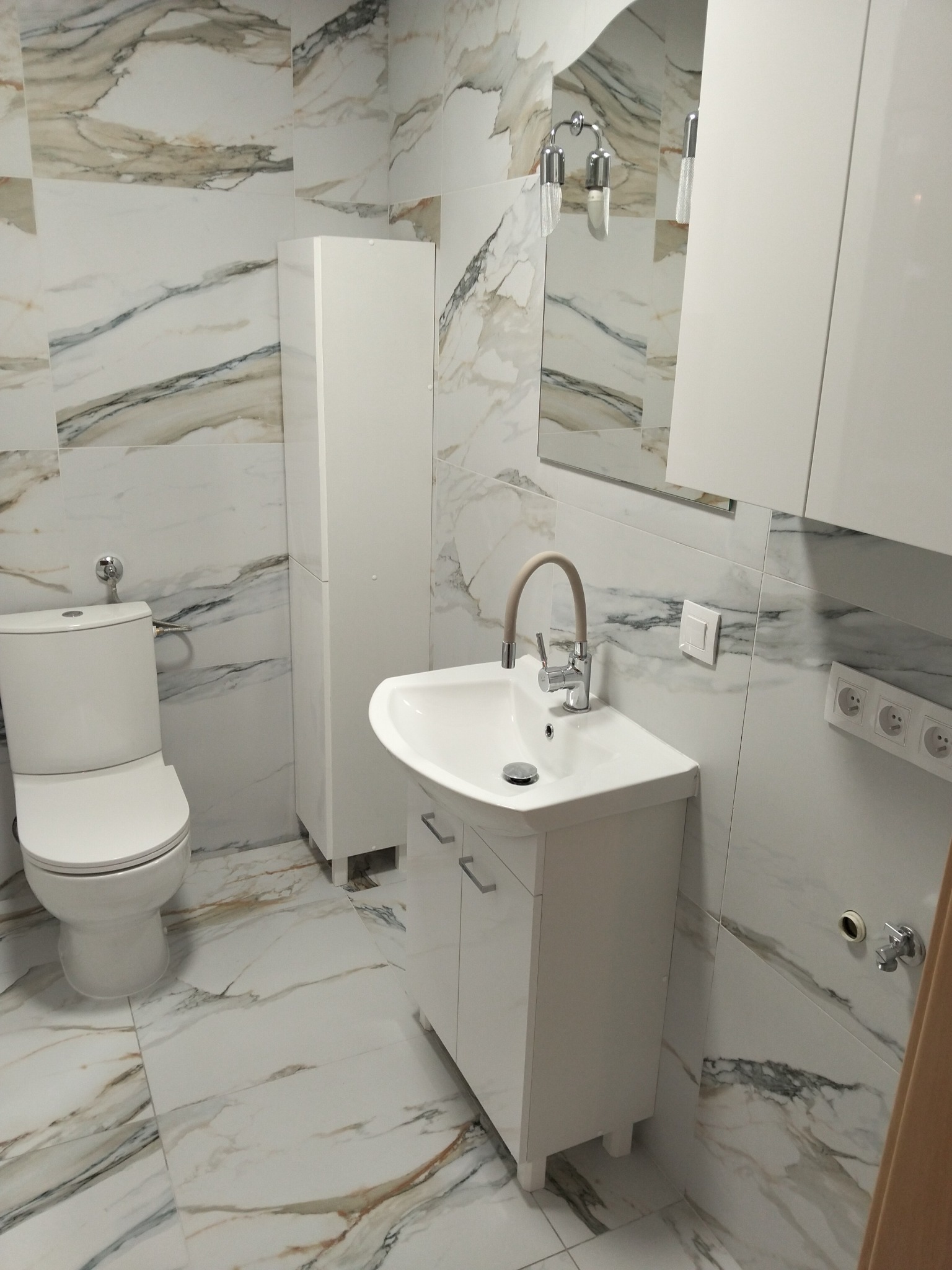 Wykończona łazienka w jasnych marmurkowych płytkach. Biała toaleta, szafka pod umywalką i słupek. Bateria z beżowym wężykiem. Lustro z oświetleniem.