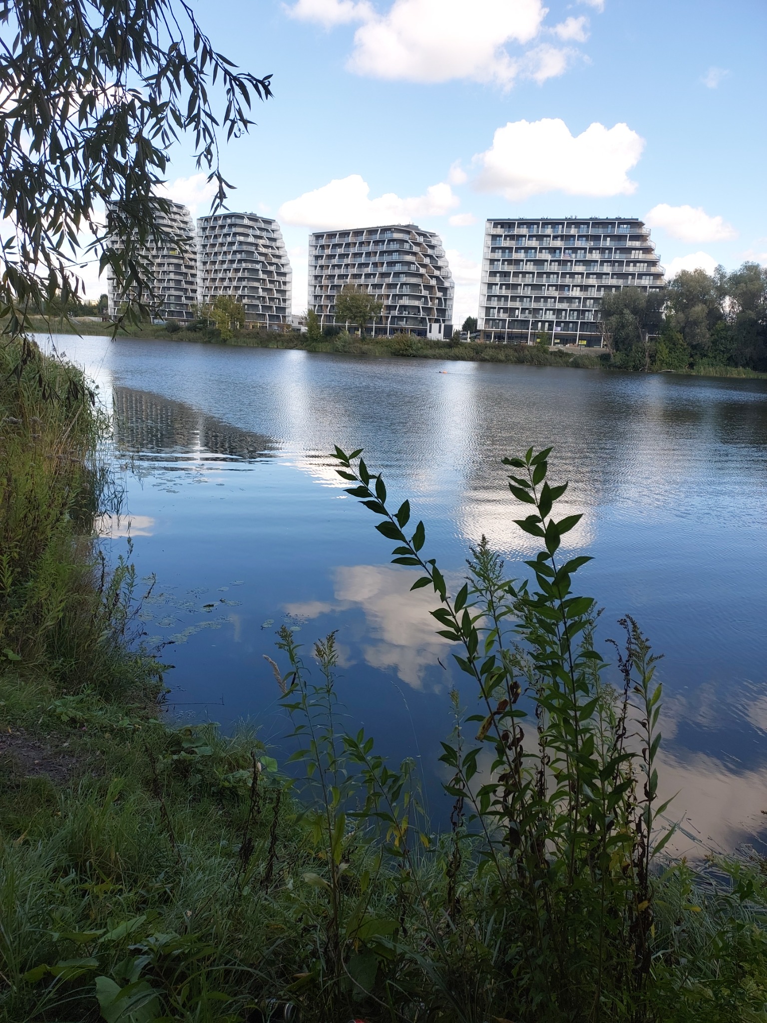 Trzy nowoczesne budynki mieszkalne z balkonami, odbijające się w spokojnej tafli wody jeziora, otoczone zielenią i błękitnym niebem z chmurami.