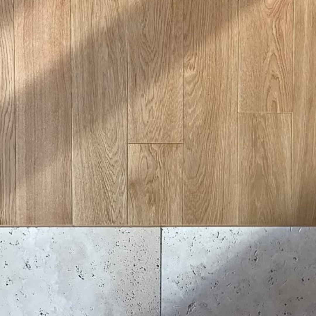 Jasne, drewniane panele podłogowe ułożone pionowo, kontrastujące z jasnymi, porowatymi płytkami u dołu kadru. Delikatne cienie podkreślają teksturę drewna.