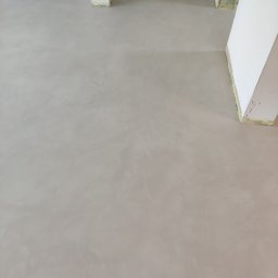 LOFTDEKOR SPÓŁKA Z OGRANICZONĄ ODPOWIEDZIALNOŚCIĄ - Jasnoszara, gładka posadzka z mikrocementu w nowym wnętrzu. Widoczne fragmenty kolumn i ścian zabezpieczonych taśmą malarską. Minimalistyczne wykończenie podłogi.