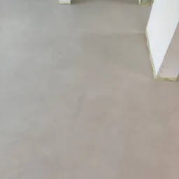 Jasnoszara, gładka posadzka z mikrocementu w nowym wnętrzu. Widoczne fragmenty kolumn i ścian zabezpieczonych taśmą malarską. Minimalistyczne wykończenie podłogi.