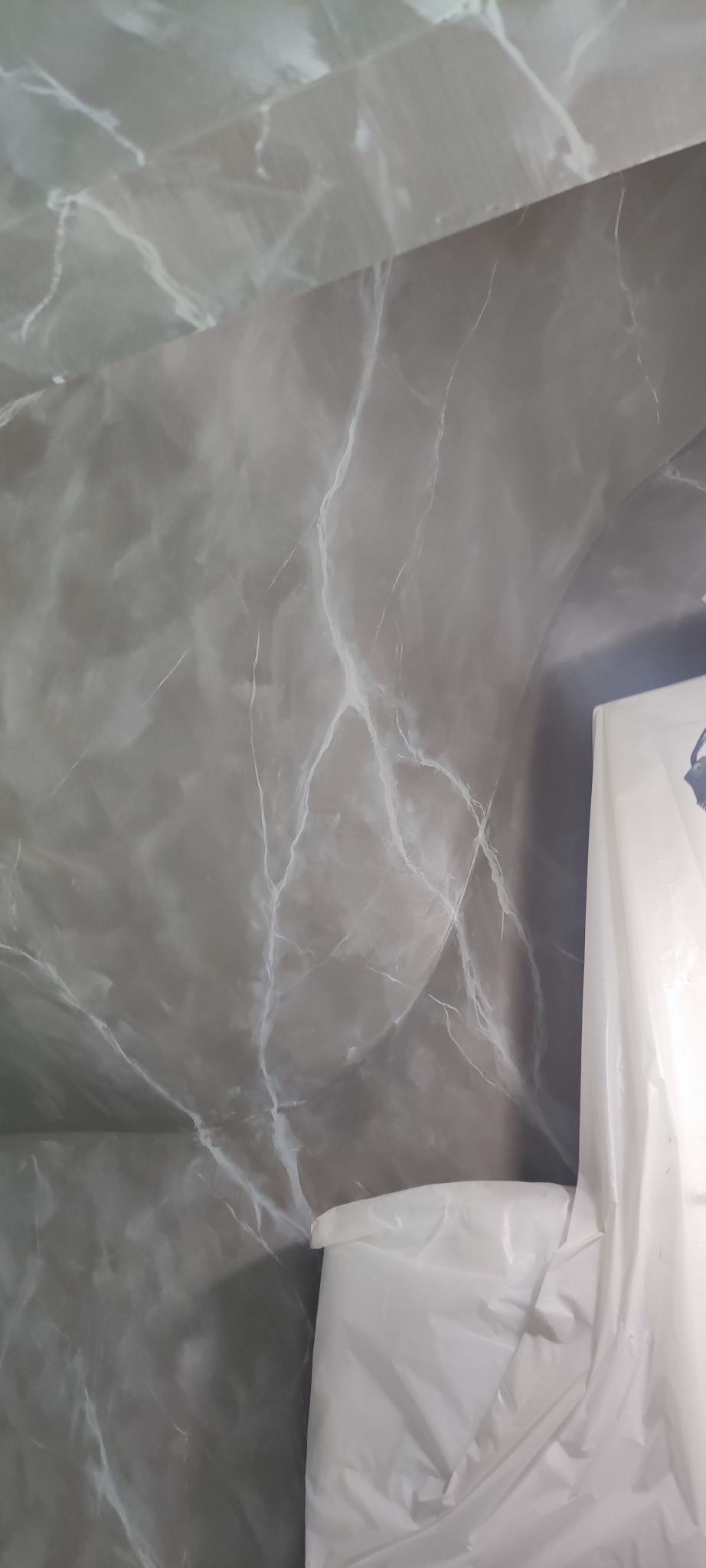 Ściana i sufit wykończone szarym mikrocementem z białymi żyłkami imitującymi marmur, częściowo zasłonięte folią ochronną podczas prac wykończeniowych.