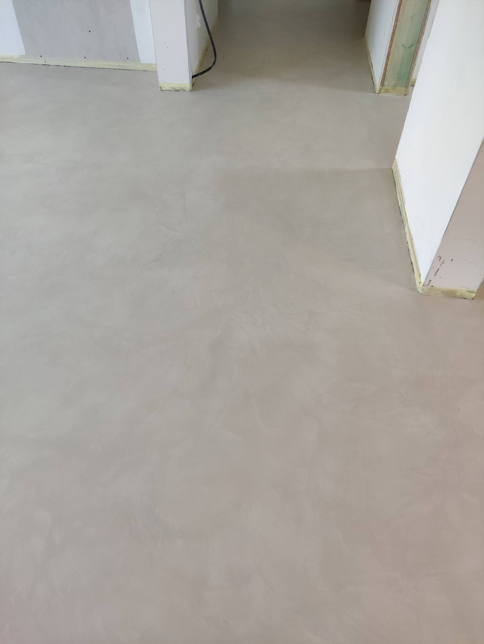 Jasnoszara, gładka posadzka z mikrocementu w nowym wnętrzu. Widoczne fragmenty kolumn i ścian zabezpieczonych taśmą malarską. Minimalistyczne wykończenie podłogi.