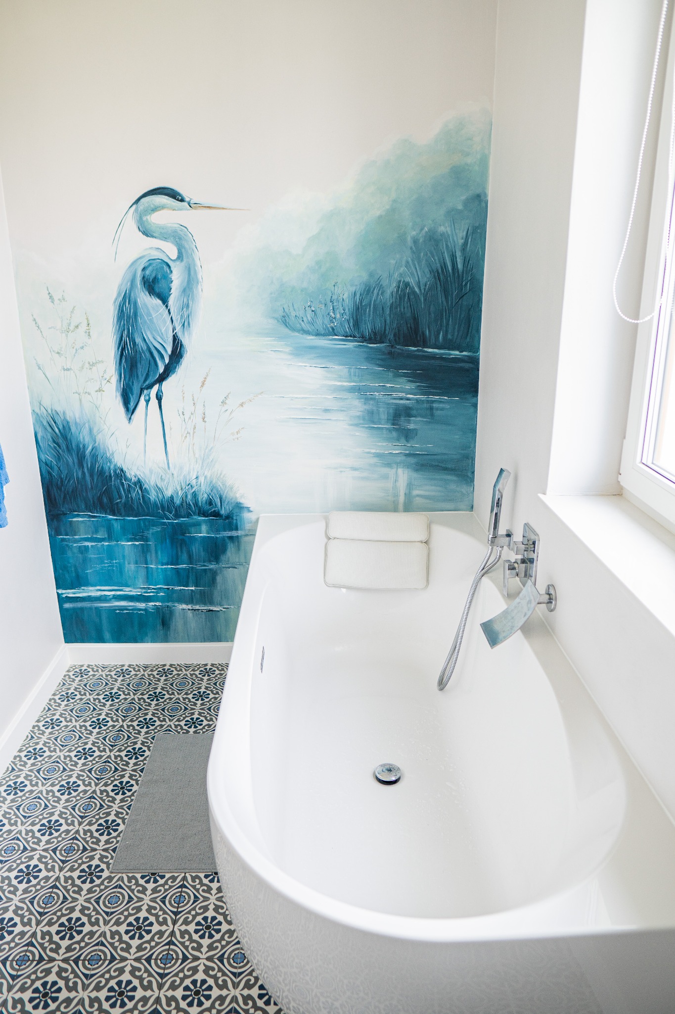 Biała wanna w łazience z dekoracyjną podłogą i malowidłem ściennym przedstawiającym czaplę nad jeziorem, widok z góry, jasne oświetlenie.