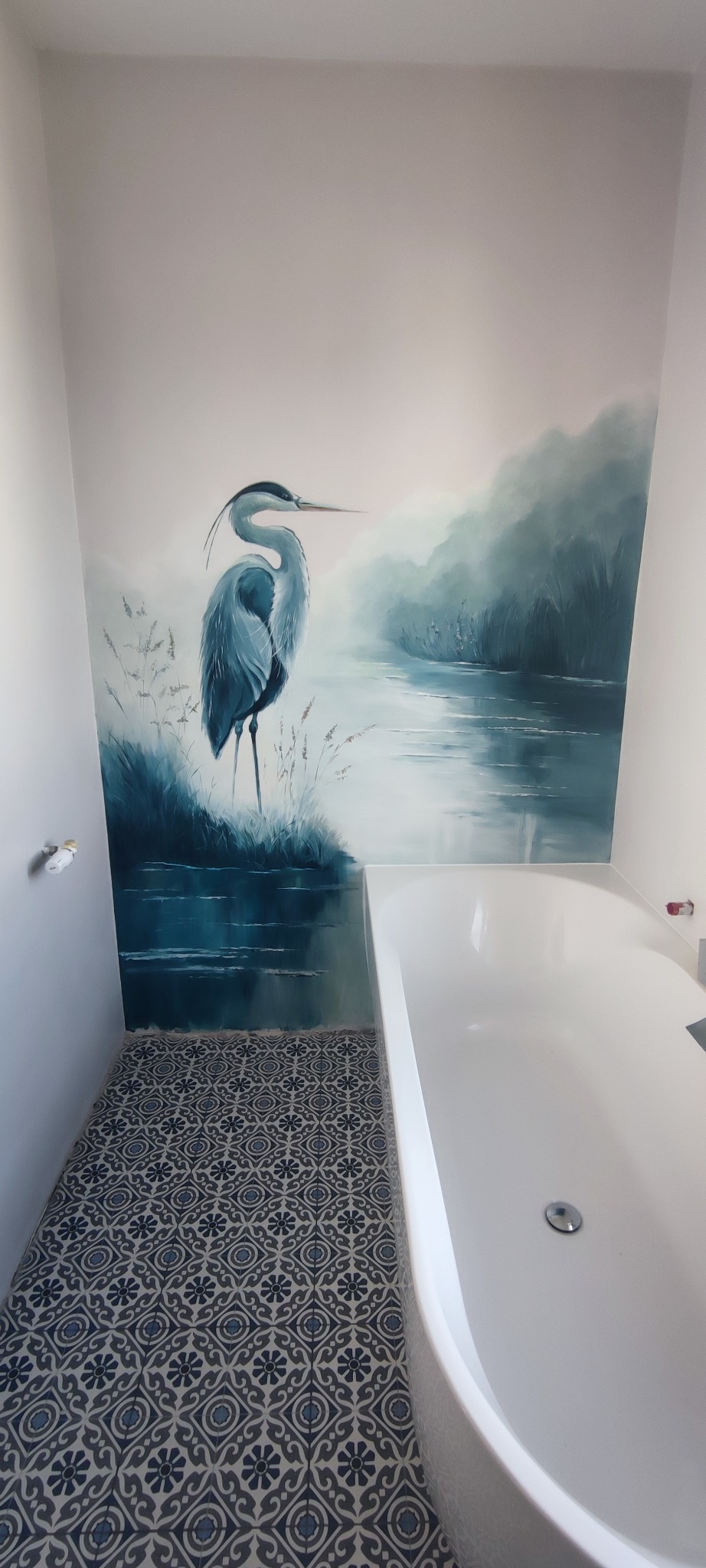 Błękitna łazienka z wanną i ścianą zdobioną malowidłem krajobrazowym z czaplą. Wzorzysta, niebiesko-szara podłoga. Ujęcie z góry, widoczny fragment instalacji.