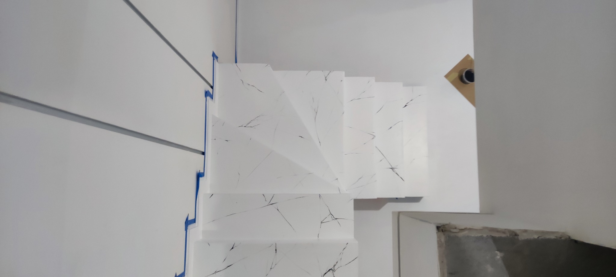 Schody wykończone białym materiałem z czarnym wzorem żyłkowania, zabezpieczone niebieską taśmą malarską przy ścianie z dekoracyjnymi listwami.
