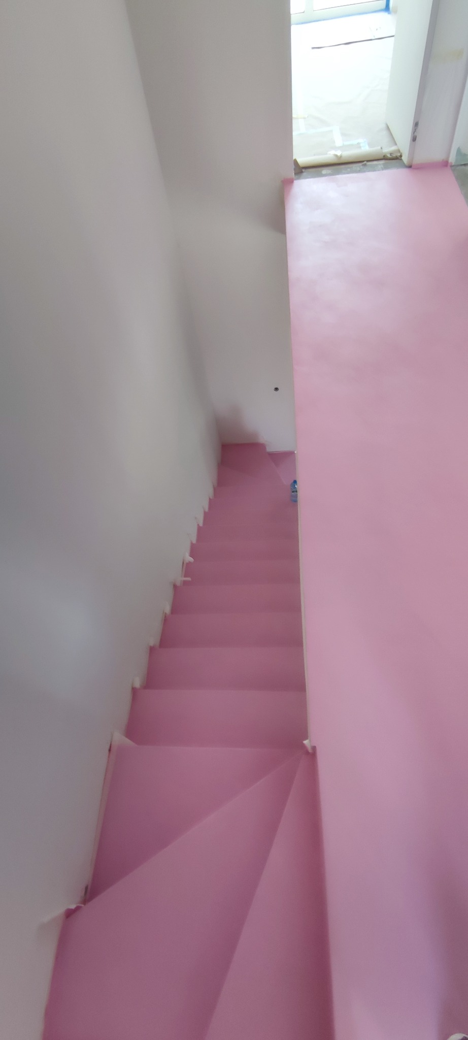Różowe schody w trakcie malowania, widok z góry. Zabezpieczona folią podłoga i ściany. Widoczne białe ściany i fragment okna w tle.