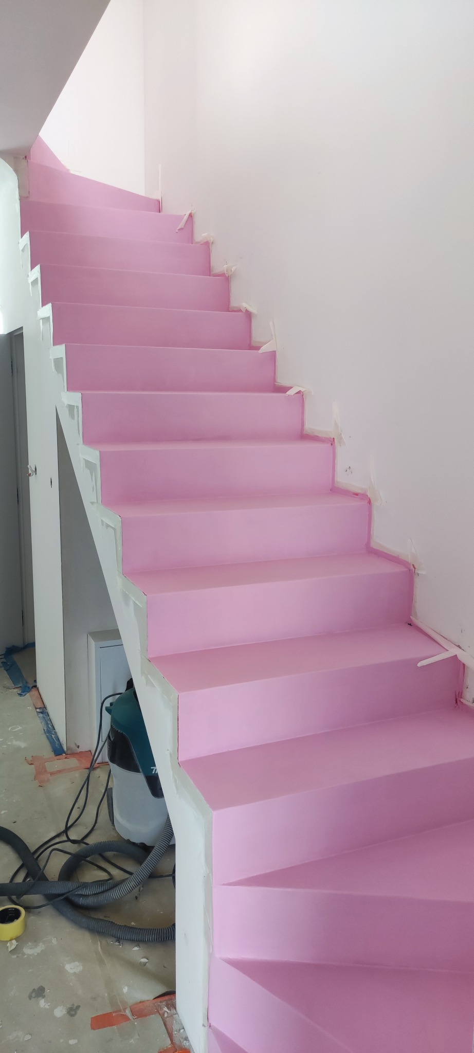 Różowe schody w trakcie malowania, zabezpieczone taśmą malarską przy ścianie. Widoczny odkurzacz przemysłowy i niedokończona podłoga.