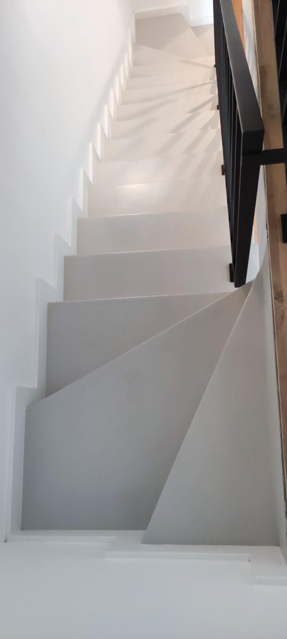 Minimalistyczne, białe schody z czarną balustradą, widziane z góry. Geometryczne kształty stopni tworzą ciekawy wzór. Oświetlenie podkreśla czystość form.