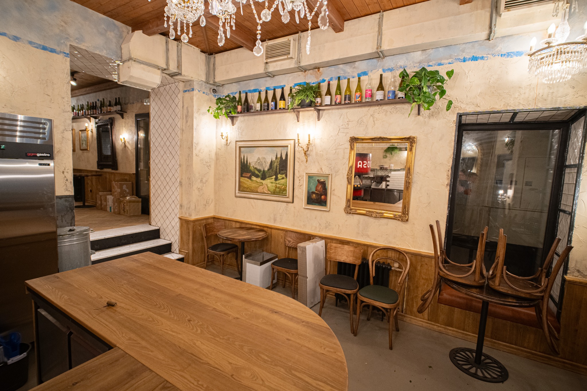 Klimatyczne wnętrze restauracji z drewnianym blatem, dekoracyjnymi obrazami, roślinami i eleganckim oświetleniem. Widoczne krzesła i elementy wystroju w stylu vintage.