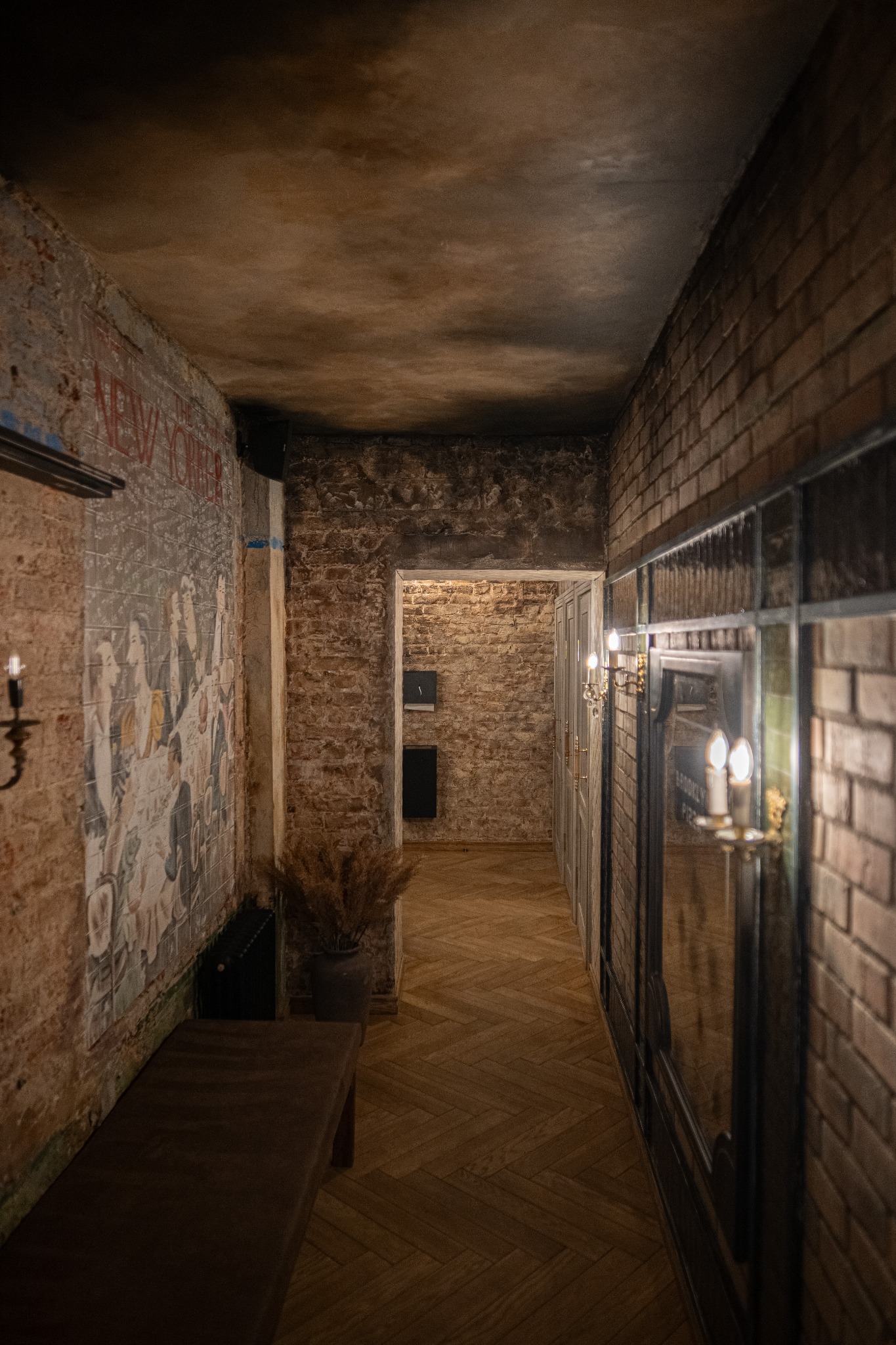 Korytarz w stylu vintage z ceglanymi ścianami, drewnianą podłogą w jodełkę i dekoracyjnym malowidłem ściennym. Klimatyczne oświetlenie świecznikami.