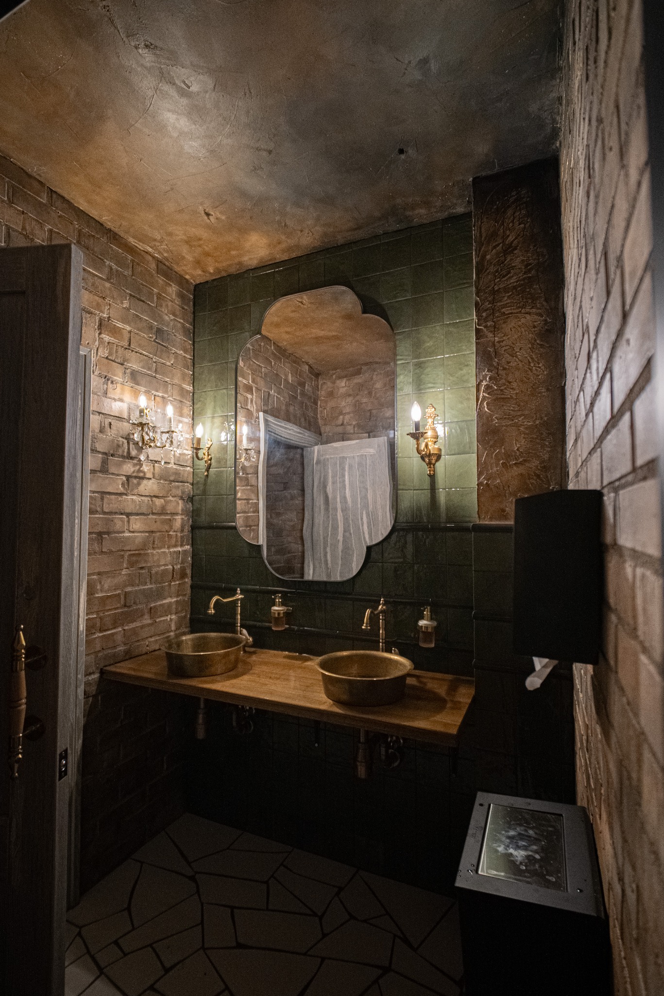 Stylowa łazienka z dwoma mosiężnymi umywalkami, ceglanymi ścianami i zielonymi płytkami. Lustro o nieregularnym kształcie odbija zasłonę prysznicową. Oświetlenie punktowe.
