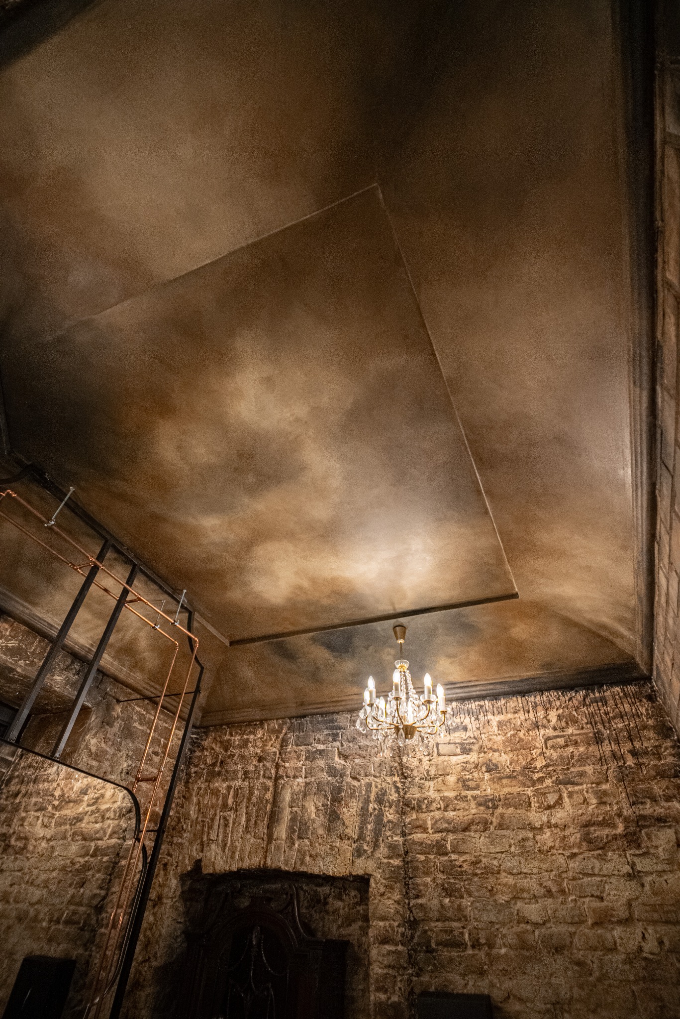 Rustykalne wnętrze z kamienną ścianą, dekoracyjnym sufitem w odcieniach brązu i kryształowym żyrandolem. Widoczne instalacje rurowe dodają industrialnego charakteru.
