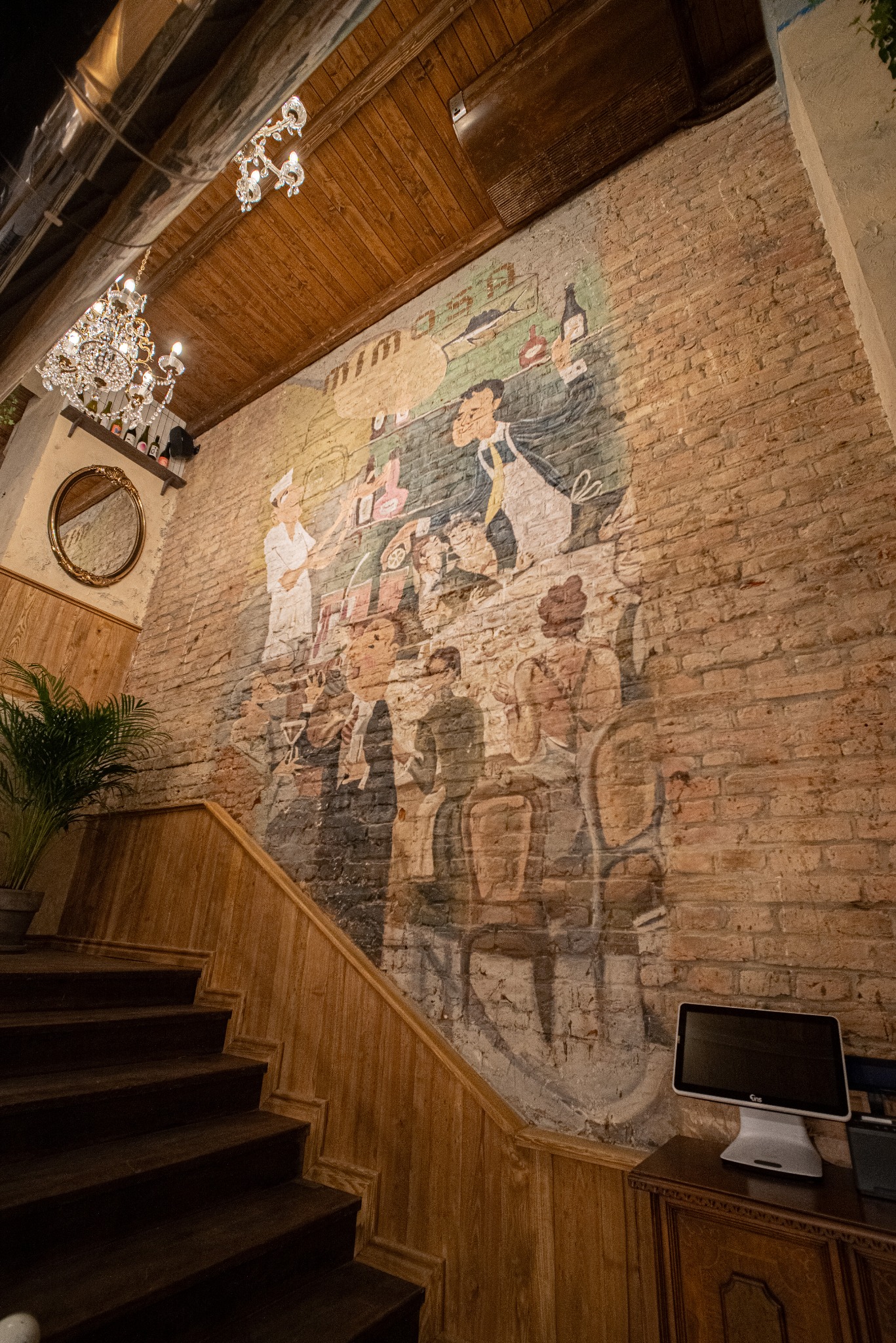 Mur z cegły z malowidłem w stylu retro przedstawiającym kucharzy i kelnerów, schody z drewnianą balustradą, kryształowy żyrandol i drewniany sufit.