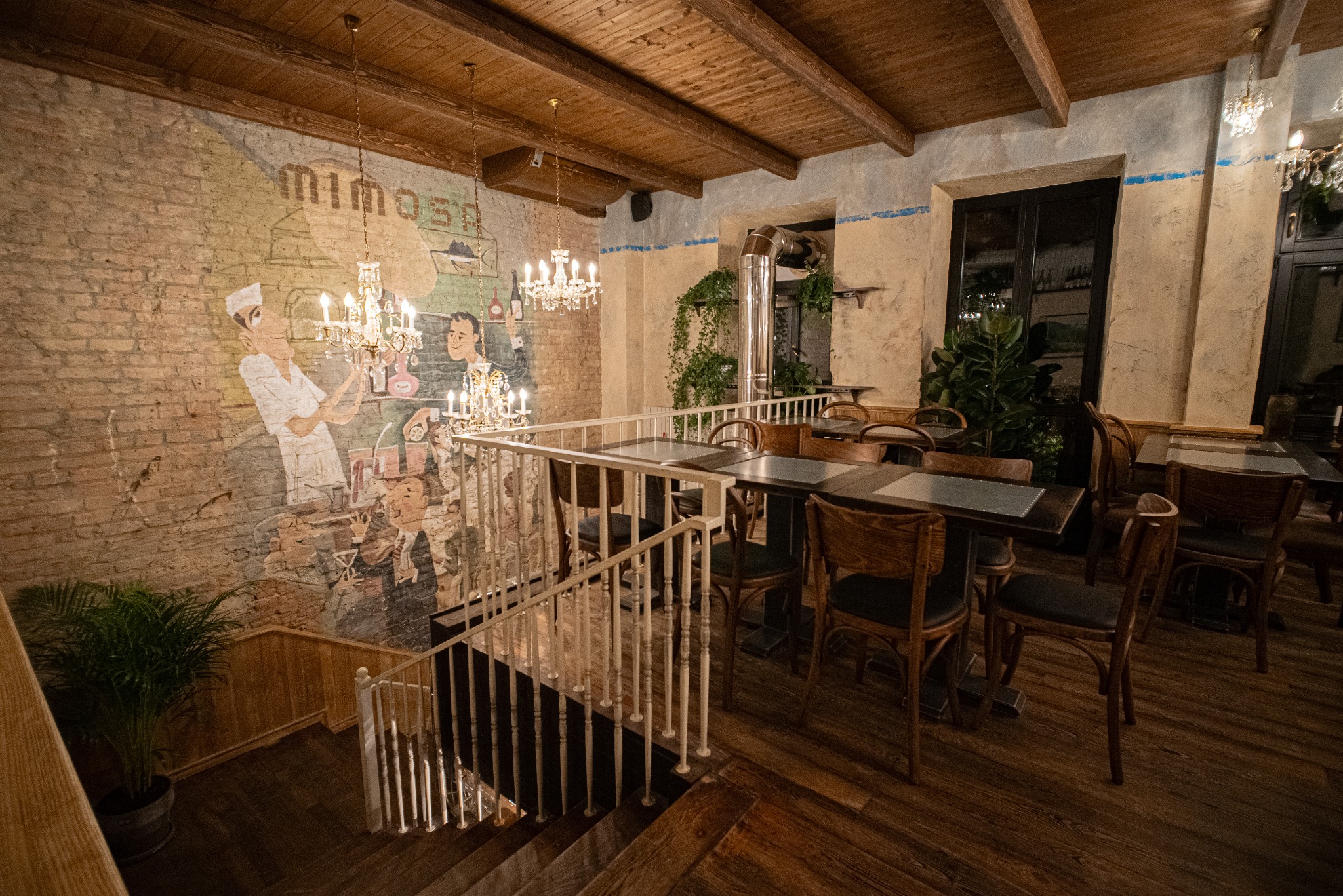 Stylowe wnętrze restauracji z muralem na ceglanej ścianie, drewnianym sufitem i schodami. Widoczne stoliki z krzesłami, rośliny i żyrandole.