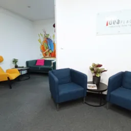 Jasne wnętrze biurowe z kolorowymi meblami: żółty fotel, niebieskie krzesła, zielona sofa. Na ścianie logo 'ideaPLACE' i mural z postacią.