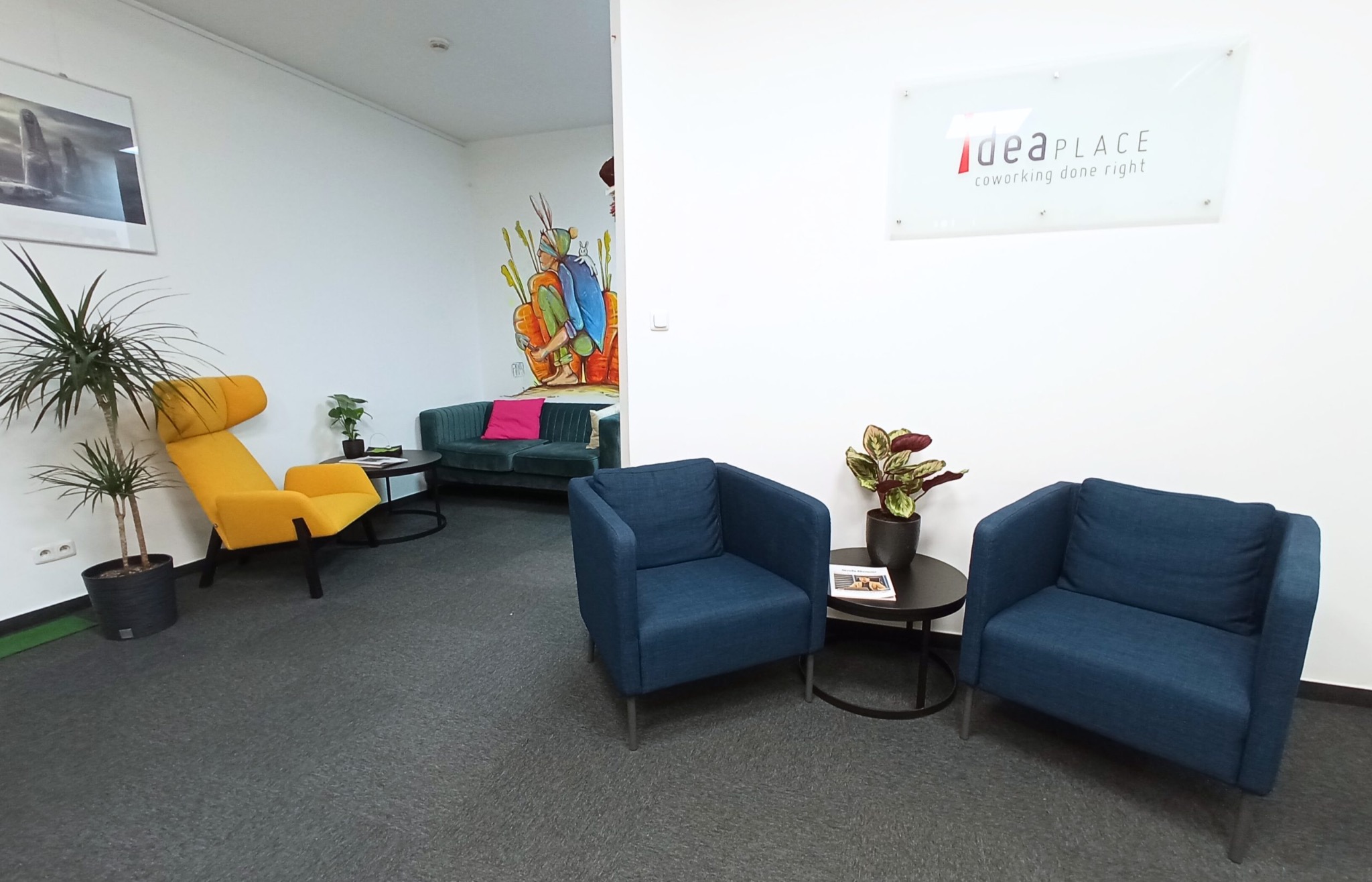 Jasne wnętrze biurowe z kolorowymi meblami: żółty fotel, niebieskie krzesła, zielona sofa. Na ścianie logo 'ideaPLACE' i mural z postacią.