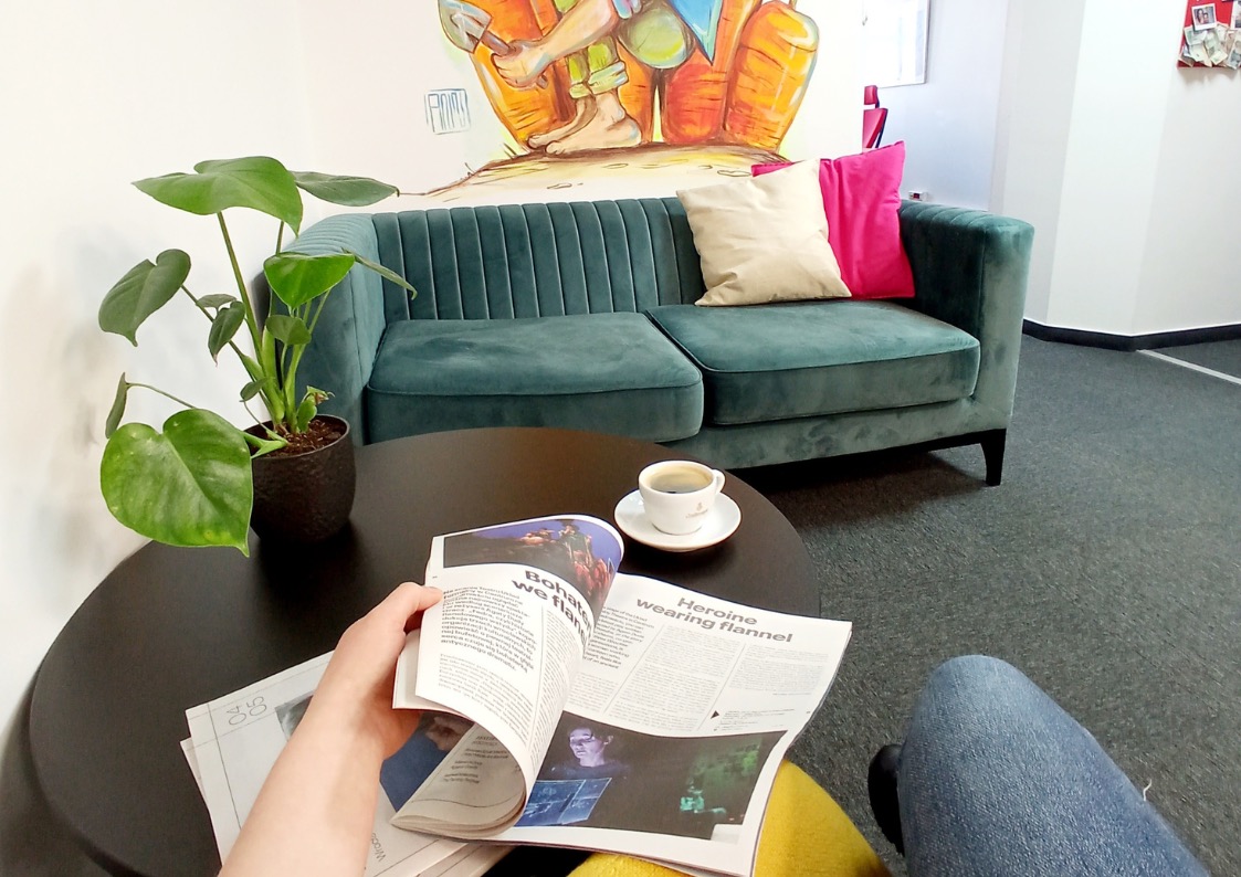 Stylowe wnętrze biura: turkusowa sofa z poduszkami, stolik z kawą i gazetą czytaną przez osobę. Na ścianie mural, roślina w doniczce. Przytulna przestrzeń do pracy.