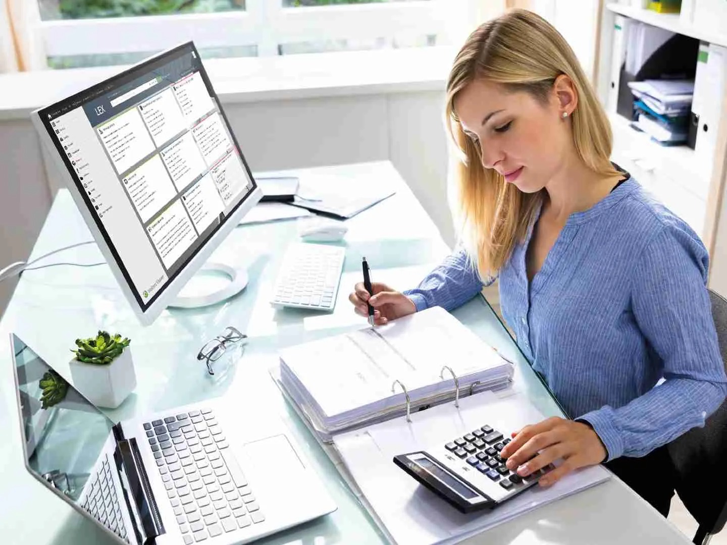 Blondynka przy biurku z komputerem, kalkulatorem i segregatorami. Kobieta w błękitnej koszuli, skupiona na pracy, z długopisem w dłoni.