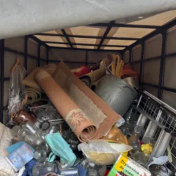 Pełny kontener z r&oacute;żnorodnymi rzeczami: dywany, słoiki, książki i sprzęty kuchenne, przygotowany do wywozu podczas opr&oacute;żniania mieszkania.