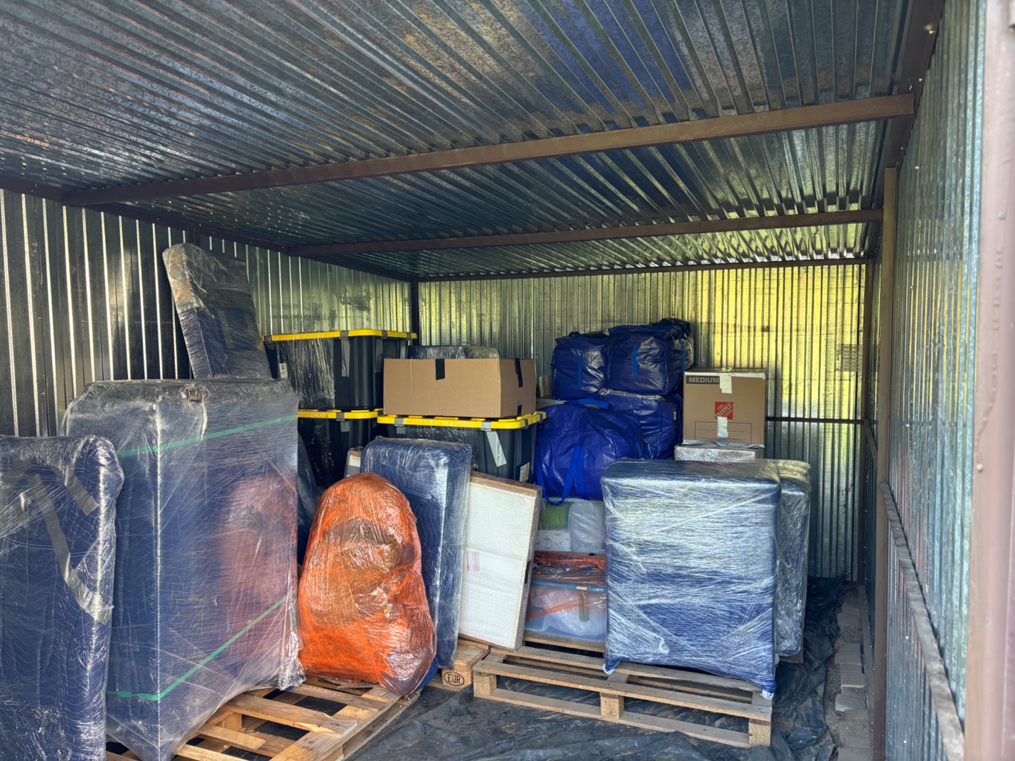 Zapakowane meble i przedmioty w magazynie z blachy falistej, przygotowane do transportu lub przechowywania. Palety, folia stretch, kartony i torby.