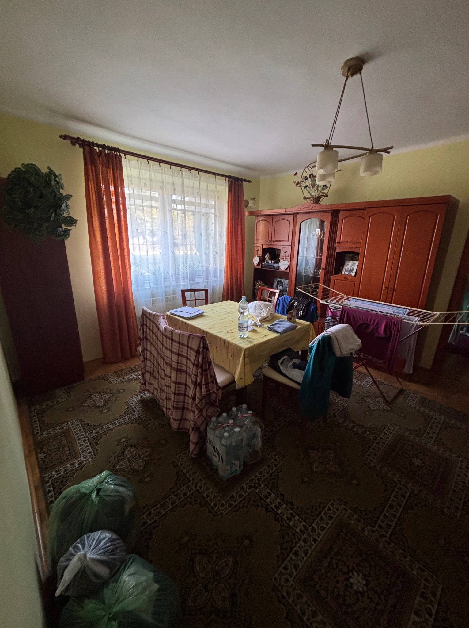 Pokój przed opróżnianiem: stół z butelką wody, krzesła z ubraniami, worki na podłodze, meblościanka, suszarka na pranie. Widok ogólny wnętrza.