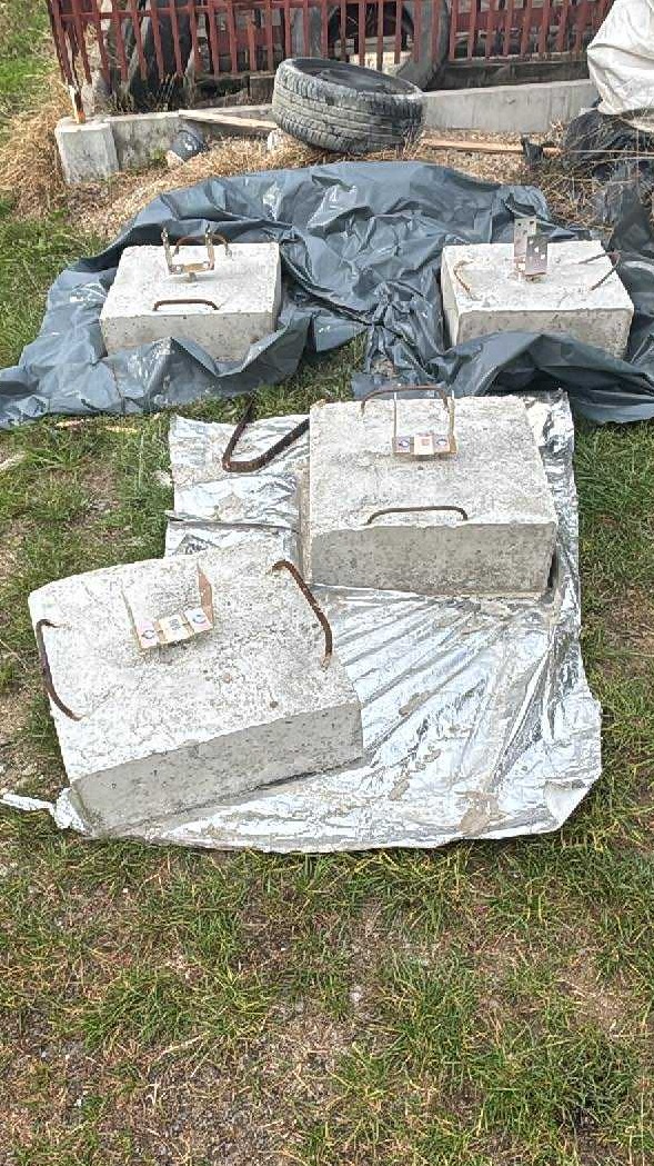 Betonowe bloki z uchwytami na folii w ogrodzie, prawdopodobnie pozostałość po demontażu konstrukcji, wskazują na potrzebę uprzątnięcia terenu.