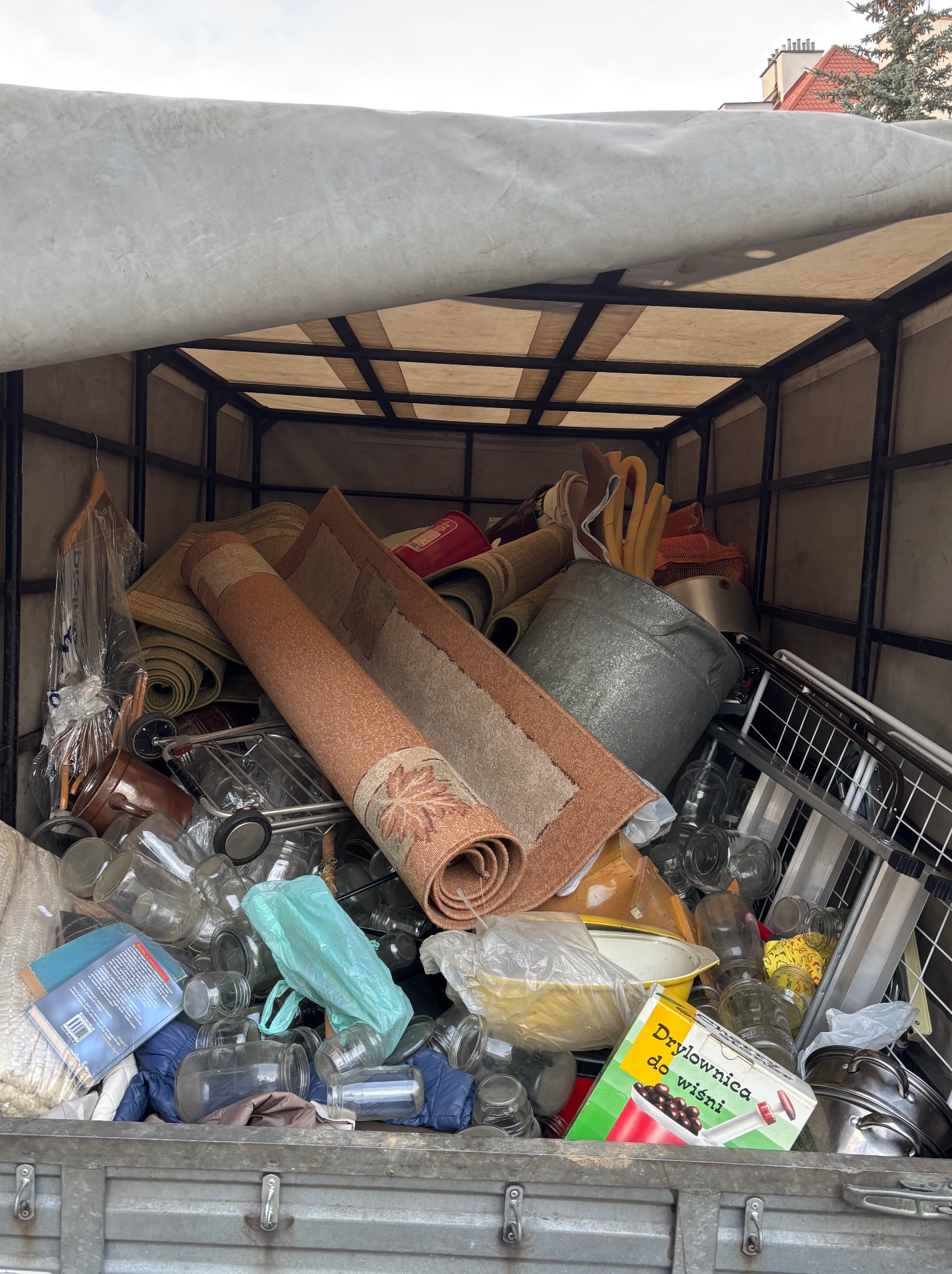 Pełny kontener z różnorodnymi rzeczami: dywany, słoiki, książki i sprzęty kuchenne, przygotowany do wywozu podczas opróżniania mieszkania.