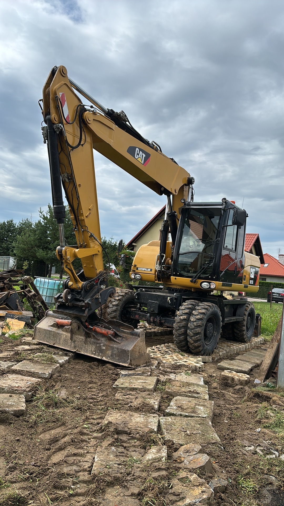 Żółta koparka kołowa CAT na placu budowy, z widocznym łyżką i fragmentami kostki brukowej. W tle dom z czerwoną dachówką i pochmurne niebo.
