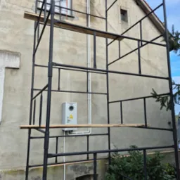 Szafka elektryczna na elewacji budynku w trakcie prac, widoczna z poziomu rusztowania. Biała rura instalacyjna. Stara, szara elewacja budynku.