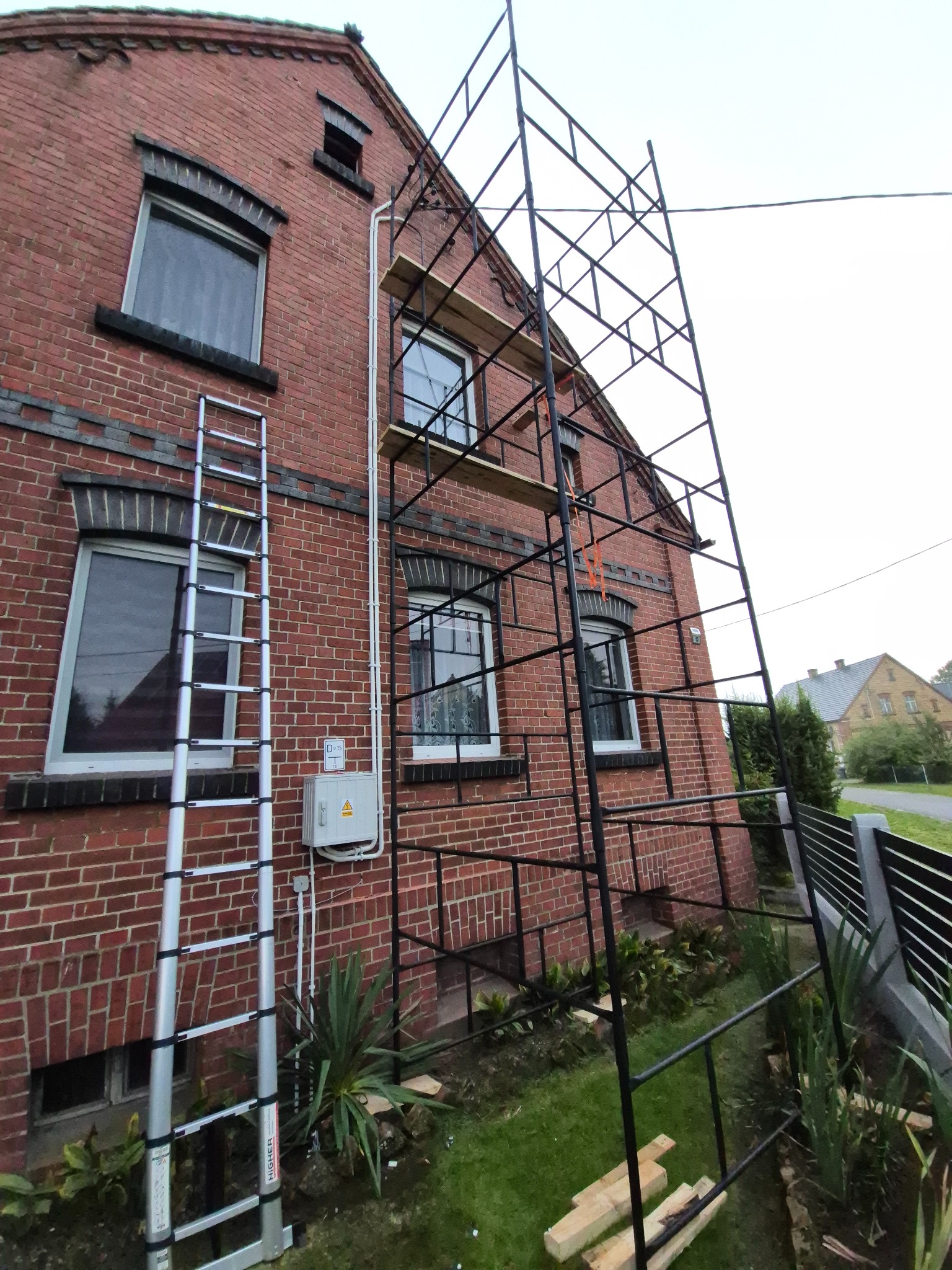 Ceglany dom z rusztowaniem i drabiną. Widoczna skrzynka elektryczna na ścianie. Prace na wysokościach.