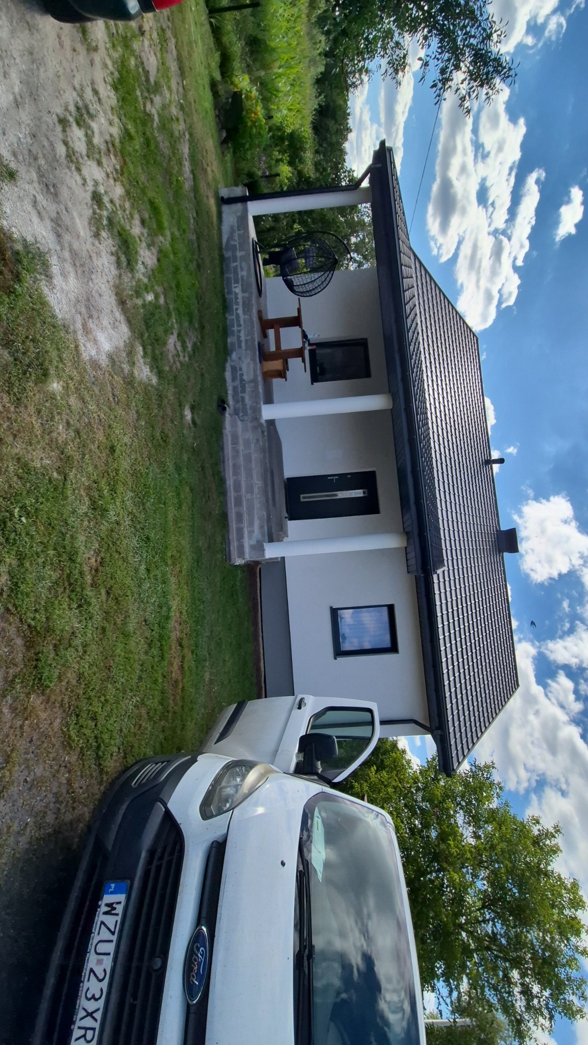 Biały, parterowy dom z ciemną dachówką, widoczny z boku, z zaparkowanym białym vanem. Na ganku huśtawka typu kokon i drewniany stolik. Błękitne niebo z chmurami w tle.