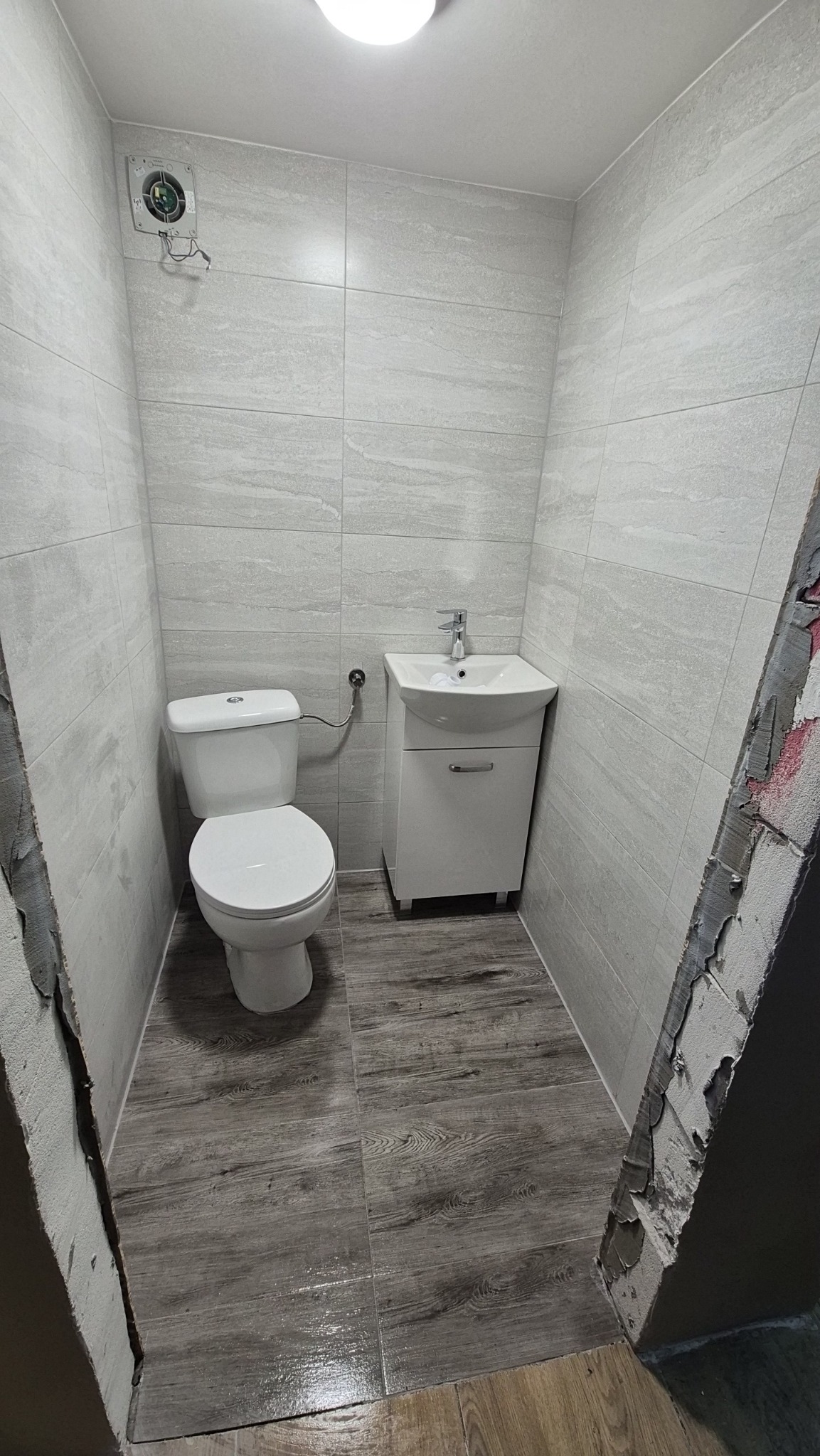 Wykończona, mała łazienka z szarymi płytkami imitującymi kamień na ścianach i drewnopodobną podłogą. Widoczna toaleta, umywalka z szafką i wentylator.