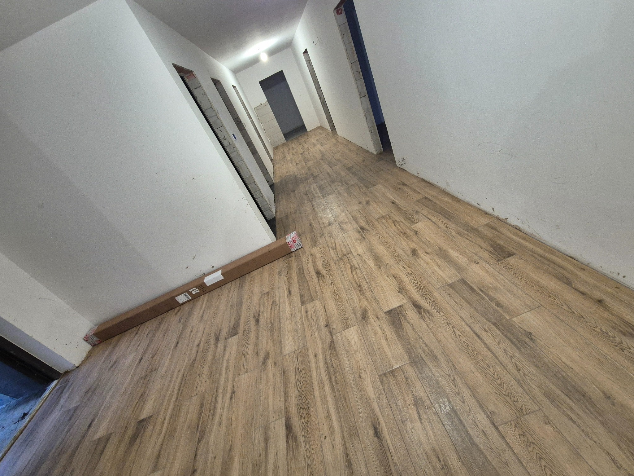 Korytarz w trakcie remontu z ułożonymi panelami podłogowymi o wzorze drewna. Na podłodze leży karton z panelami. Ściany białe, surowe otwory drzwiowe.