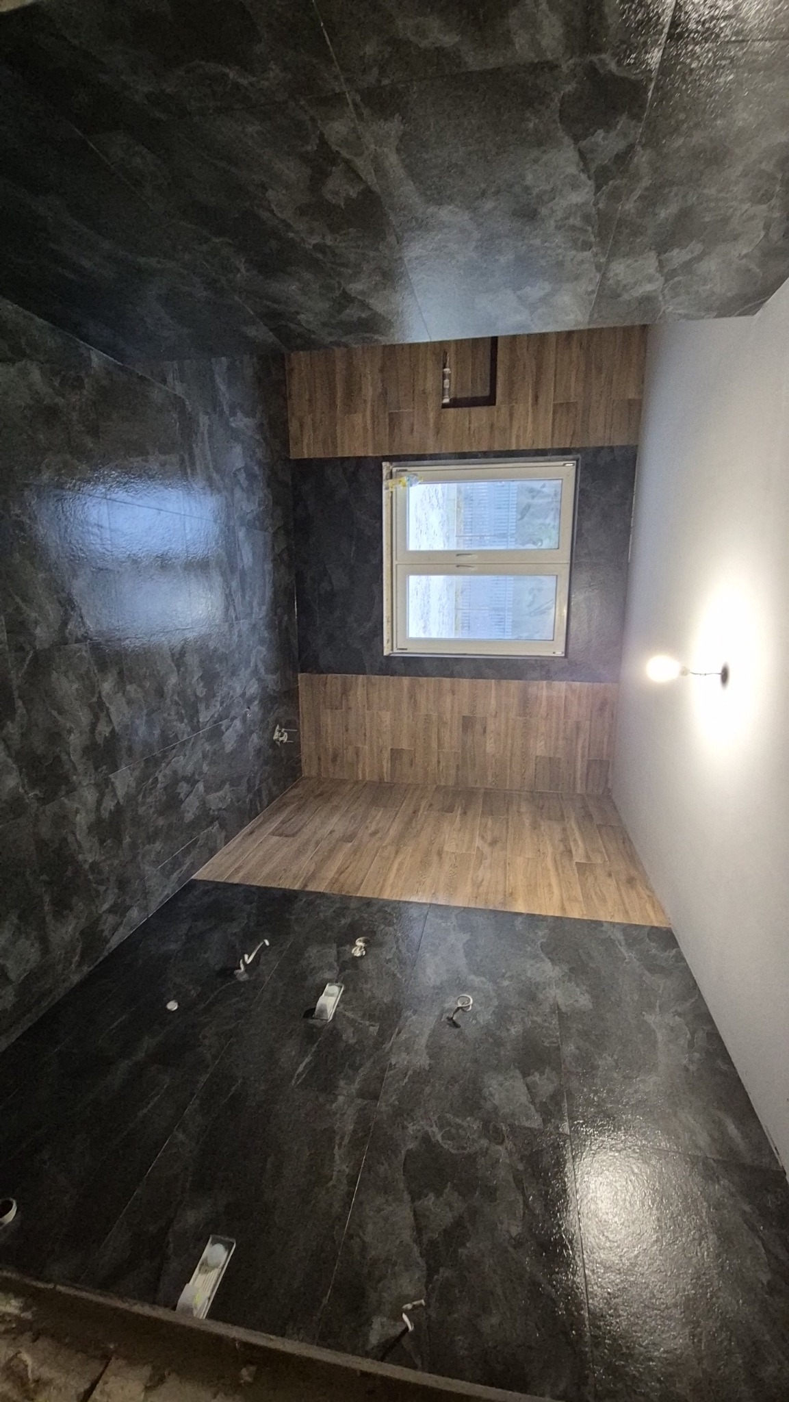Nowoczesna łazienka po remoncie: ciemne, marmurkowe płytki na ścianach i podłodze, drewnopodobne panele na górze ścian, okno z widokiem na zewnątrz, przygotowane punkty instalacji.